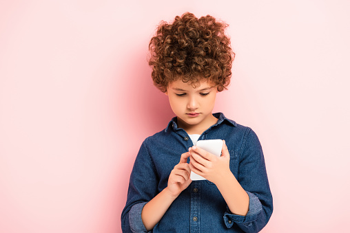 curly kid in denim shirt using smartphone on pink