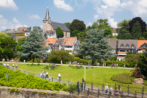 People on promenade and Essen Kettwig in spring season. View over river Ruhr from bridge towards old historical center with church and buildings around and below. People are walking along promenade at Ruhr