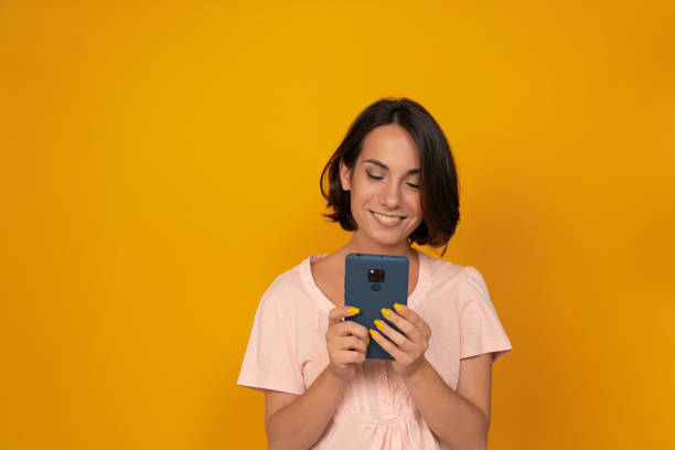 istock Young smiling woman scrolls or types in mobile phone. Isolated on yellow background. Copy space 1281792884