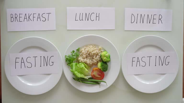 Hands of a woman put a plate with brown rice and vegetables under the title lunch, and under the title breakfast and dinner she puts on empty plates titles fasting. Interval fasting concept. Skipping meals