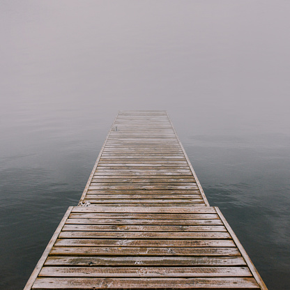 Wooden pier that goes into the fog. Empty mysterious bridge on the river made of planks. Quiet foggy landscape. Dramatic atmosphere. The concept of solitude and serene mood.