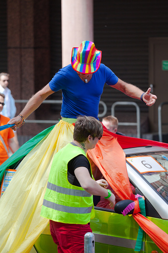 Man getting decorated at CSD parade with rainbow colored curtains. Man is standing on a convertible car and is wearing a multicolored hat