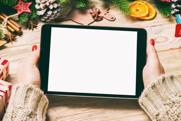 Photo of Top view of female hands holding a tablet on wooden Christmas background made of fir tree and festive decorations. New year holiday concept. Mockup