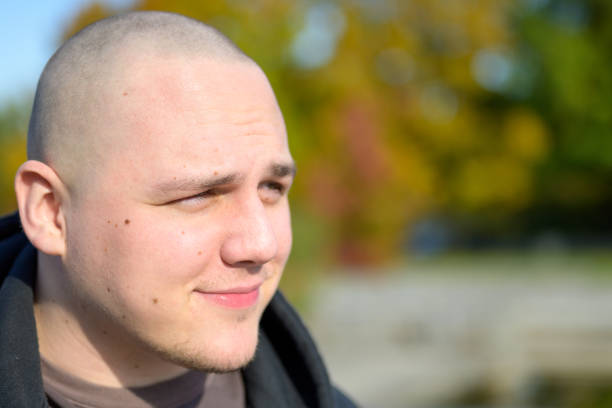 Thoughtful young man with a quiet smile squinting against the warm autumn sunshine as he poses outdoors in a garden in a close up head shot looking to the side