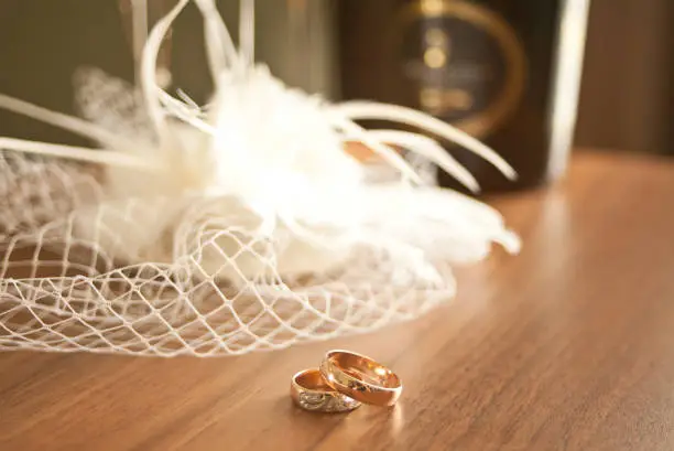 Photo of wedding rings symbol attributes lie on a table with hat or veil, glasses and bottles of champagne.