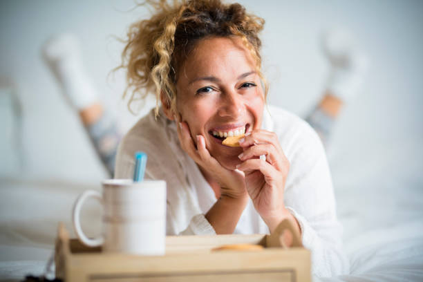 retrato de mulher adulta linda comendo biscoito no café da manhã no quarto - casa ou hotel acorda dia com pessoas bonitas do sexo feminino deitada com biscoito e chá - cookie women eating beautiful - fotografias e filmes do acervo