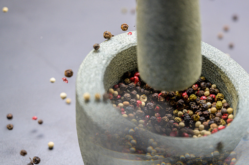Close-up view of various kinds of pepper in stone mortar. Grinding pepper in mortar with pestle. Several peppercorns falling out from mortar.