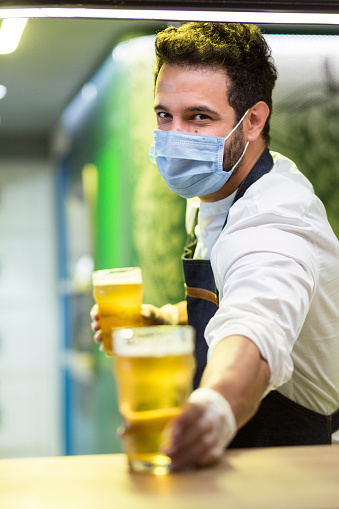 Bartender man of Latino ethnicity between 25-35 years old is serving beer with all the biosecurity measures due to the COVID-19 pandemic