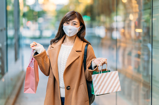 A woman is holding shopping bags and smiling for the camera with a protective face mask for illness prevention.