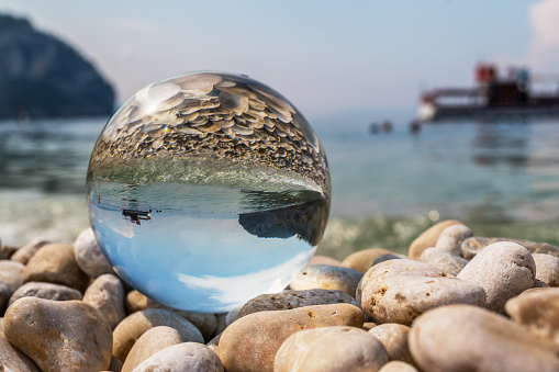 Crystal ball on pebbles near the sea. Original upside down view and rounded perspective of the sky, sea and boat. Original and engaging picture.