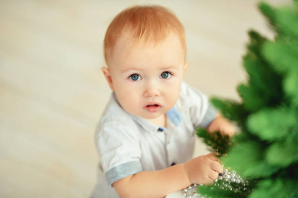 A small blond boy in blue jeans and a white shirt stands at a table with a Christmas tree. Horizontal photography