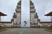 Beautiful Girl’s Full-Length Portrait Near Gates Of Heaven In Pura Lempuyang Temple In Bali, Indonesia. Happy Young Woman Doing Back Band Jump Near Ancient Architecture In Asia.
