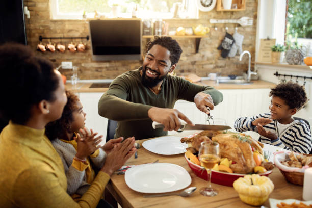 Happy black father carving roasted turkey during Thanksgiving lunch at dining table.
