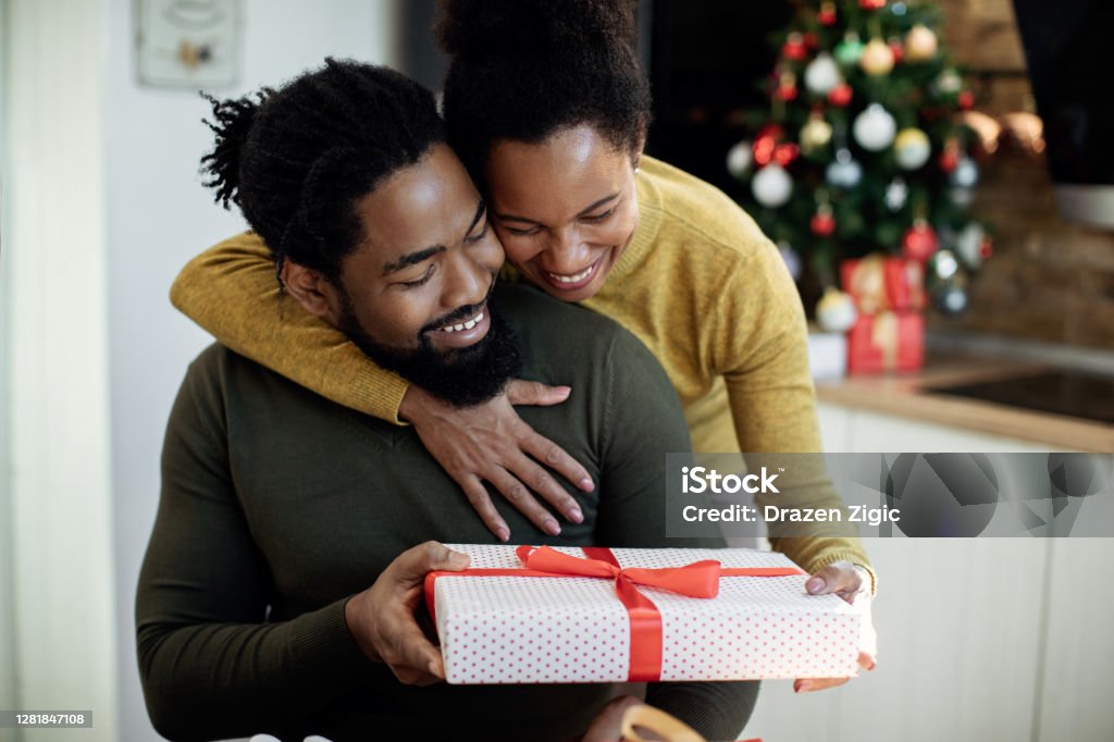 Happy African American woman surprising her husband with Christmas present. Happy black woman embracing her husband and giving his a gift on Christmas day at home. Christmas Present Stock Photo
