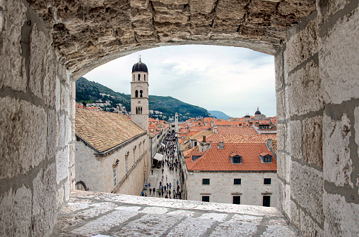 View from stone window of Dubrovnik city center, old tower and crowd. Crowded touristic narrow street in old town, View of red rooftops and tower.