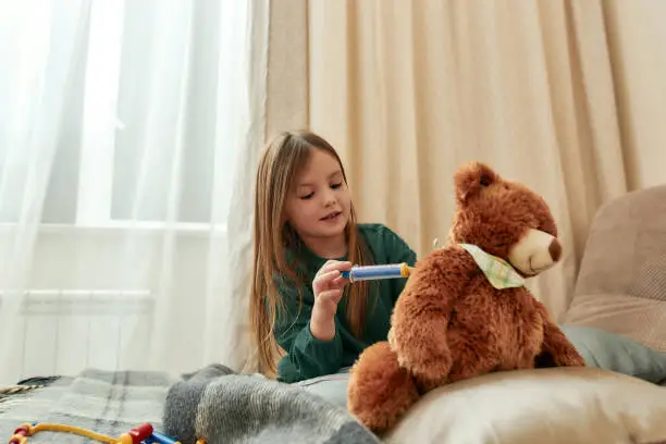 A small girl making a toy injection in a teddybear's back playing doctor sitting on a sofa in a bright guestroom being alone at home. Childrens leisure activities