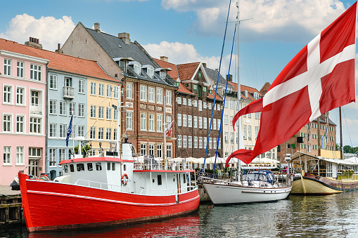 Nyhavn a 17th century harbour in Copenhagen with typical colorful houses and boats with national flag of Denmark on the first ground.