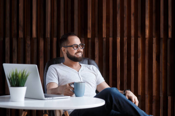 Young successful bearded businessman freelancer in office coworking. Happy man programmer developer in a white t shirt smiling and drinking coffee at the Desk with a laptop