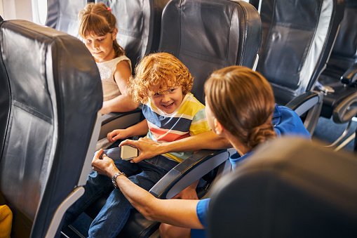 Kid sitting on an aisle seat laughing joyfully with an air hostess putting a safety belt on him