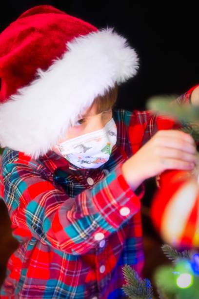 istock Serious Serious pensive Little boy wearing a Santa hat and a protective face mask decorating a Christmas Tree 1281880095