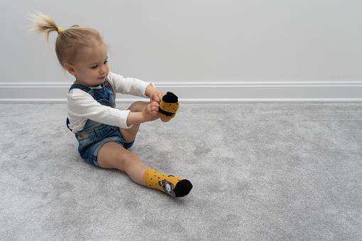 Cute caucasian baby girl learning to put on socks at home, on floor, white wall behind. Casual wear, indoors.