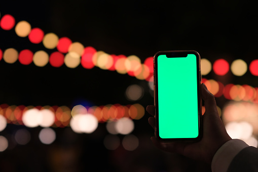 close up one hand showing green screen smart phone in amusement park at night. Defocused holiday lights