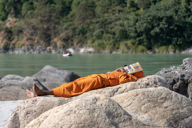 hindu sadhu heiliger mann, schläft am ufer bedeckt seinen kopf mit einer zeitung an einem sonnigen tag in der nähe des ganges flusswasser in rishikesh, indien, aus nächster nähe - hinduism outdoors horizontal close up stock-fotos und bilder