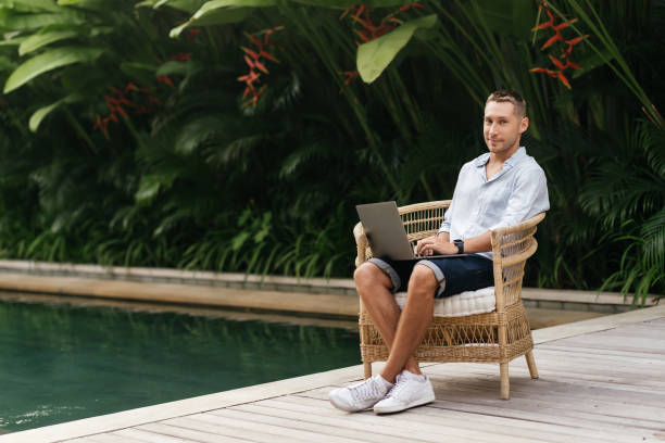 istock Young man using a laptop computer in a garden with a swimming pool. 1281947270