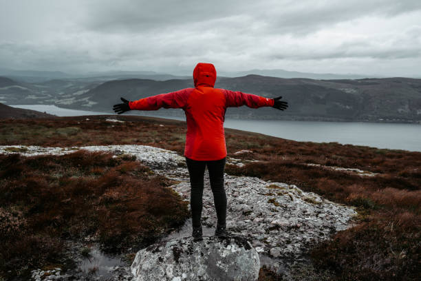 The woman in the red jacket stands with her arms outstretched, below Loch Ness and the hills covered with heather.