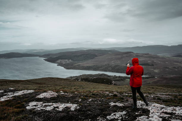A tourist in a red jacket admires Loch Ness and hills covered with heather.