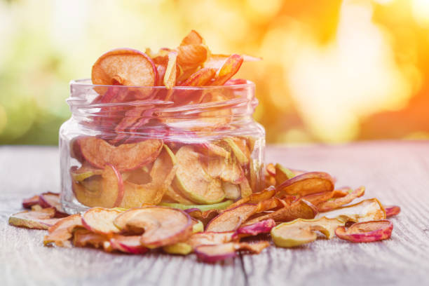 homemade apple chips. dried fruit. healthy snack in a glass jar and on a wooden surface with green blurry background and sunlight - dried apple imagens e fotografias de stock