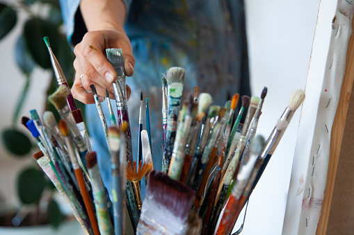 Woman artist hand choosing and picking oil paintbrushes near canvas in workshop. Art and craft supplies concept. Stack of mixed sizes of painting brushes.