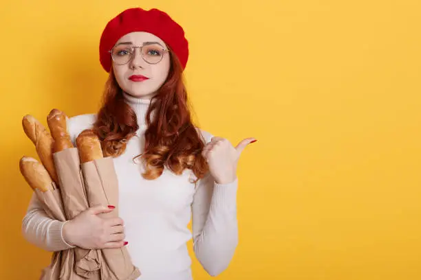 Photo of Young beautiful upset woman holding bag of fresh healthy bread over yellow background,red haired sad girl indicating aside, looks worried and nervous, lady wearing beret, white shirt and glasses.