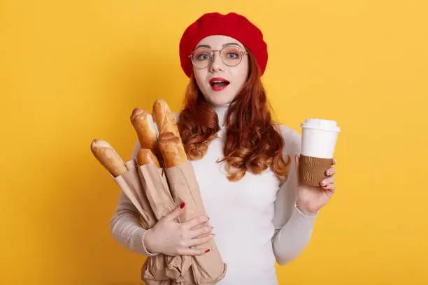 Photo of Portrait of astonished woman wearing beret, white shirt and glasses, holding paper bags with fresh baguettes and coffee to go, female with opened mouth and surprised facial expression.