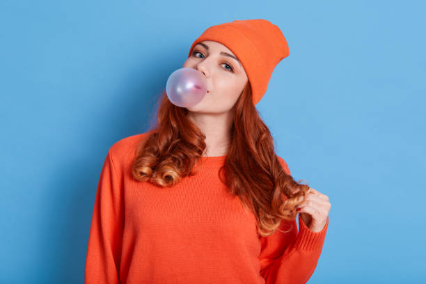 istock Beautiful young woman blowing bubblegum while posing isolated over blue background, wearing casual clothing and hat, having red hair, attractive girl with wavy hair. 1281977315