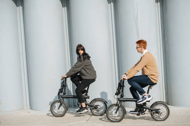 Young man and woman riding ebikes, looking at each other. On a sunny autumn day Young man and woman riding ebikes, looking at each other. On a sunny autumn day. Big pillars in a background. photo of thick woman in glasses stock pictures, royalty-free photos & images