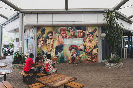 Port Moresby, Papua New Guinea - June 15, 2019: People sit out side the terminal near a beautiful mural at Jacksons International Airport in Port Moresby, Papua New Guinea