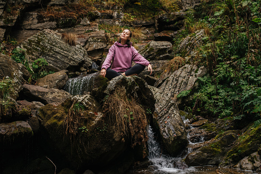 Young woman sitting barefoot on a big stone cross-legged, meditating, enjoying air and sound of running water. Over mountain spring with huge square boulders and autumn deciduous trees above.
