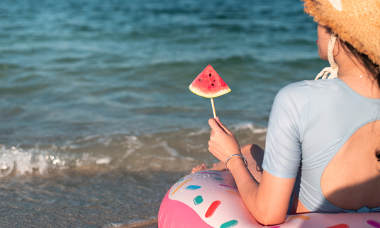 Woman eating watermelon fruit on the beach while sitting on a floating doughnut in the water. Seaside, summer vacation and leisure abstract