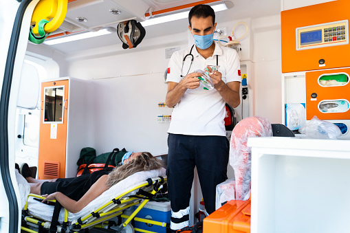 A paramedic healthcare emergency staff man wearing a medical face mask taking care of a lying down young woman patient with medical ventilator system and bag-valve masks inside an ambulance car during the coronavirus pandemic. Covid-19 infection respiratory insufficiency and artificial ventilation concept.