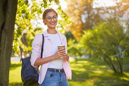 Businesswoman standing summer park Business person Outdoors Successful european caucasian woman freelancer or teacher walking outside with black backpack dressed denim jeans white shirt pink jacket