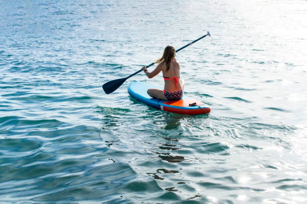 young caucasian woman from the back sitting and swimming on a blue pad on the sea. paddleboarding, activity, sup - women paddleboard bikini surfing imagens e fotografias de stock