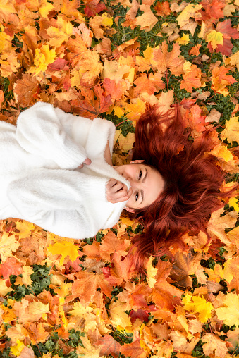 Young red hair woman smiling and lying on autumn leaves, top view. Happy beautiful girl in white sweater lying on maple leaves in autumn colors.