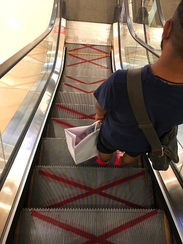 Stock photo showing the Covid-19 social distancing guidelines enforced for the use of metal escalator. Red spray painted crosses on the step help to keep people six feet apart when using the moving staircase steps.