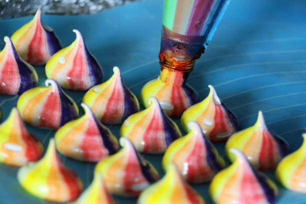 Close-up image of icing bag nozzle piping batch of miniature, homemade, circus meringue kisses on blue, silicon baking sheet, multi-coloured rainbow, raw meringue peaks ready for oven Stock photo showing close-up view of rows of raw meringue peaks being piped from icing bag nozzle on to a blue, silicon baking sheet. The freshly piped rainbow coloured, circus meringue kisses are coloured with purple, red, orange and yellow food colouring. food coloring stock pictures, royalty-free photos & images