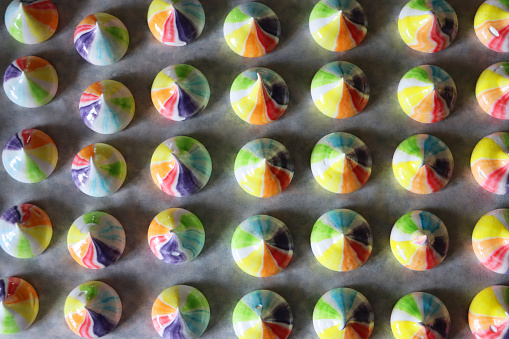 Stock photo showing close-up view of greaseproof baking parchment containing rows of multi-coloured, piped raw meringue peaks. The freshly piped rainbow coloured, circus meringue kisses are coloured with blue, green, orange, red, purple and yellow food colouring.