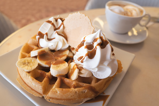 Banana, caramel and whipped ice cream waffle with a wafer next to a n espresso coffee with ice-cream in a cup and saucer.