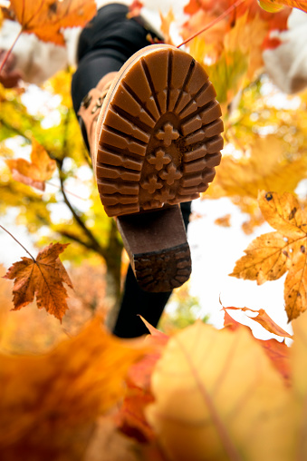 Close-up of a woman foot in boots, walking on autumn leaves. Playing with dry leaves. Autumn outdoor fun. Bottom-up view of a shoe stepping on leaves.