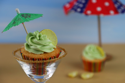 Stock photo showing close-up view of a pina colada, tropical cocktail cupcake in paper cake case sat in a martini glass against sandy beach and clear blue sky background. Afternoon tea concept.