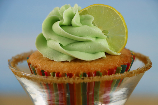 Stock photo showing close-up view of a pina colada, tropical cocktail cupcake in paper cake case sat in a martini glass against a sandy beach and clear blue sky background. Afternoon tea concept.